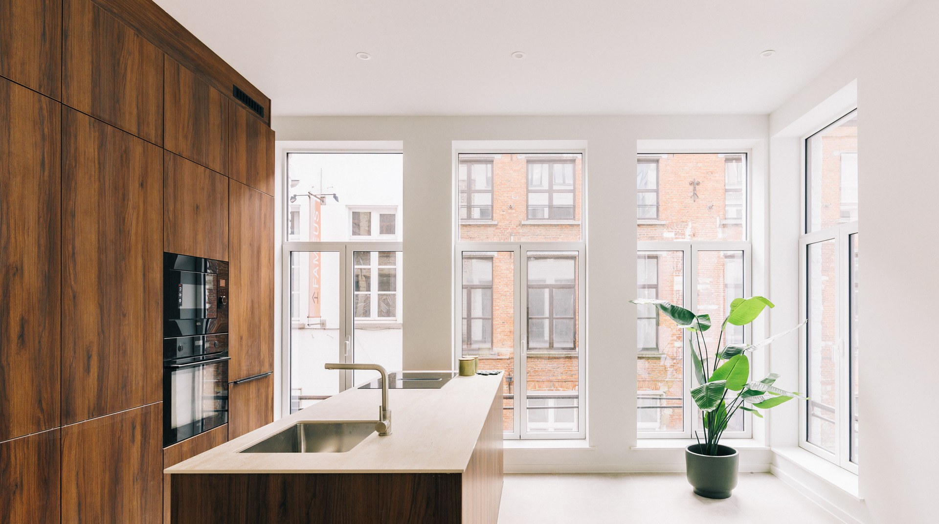 Keuken met een kastenwand in notelaar tot het plafond, ingebouwde ovens en een eiland met een blad in keramische travertijn, met hoge ramen op Antwerpse gevels