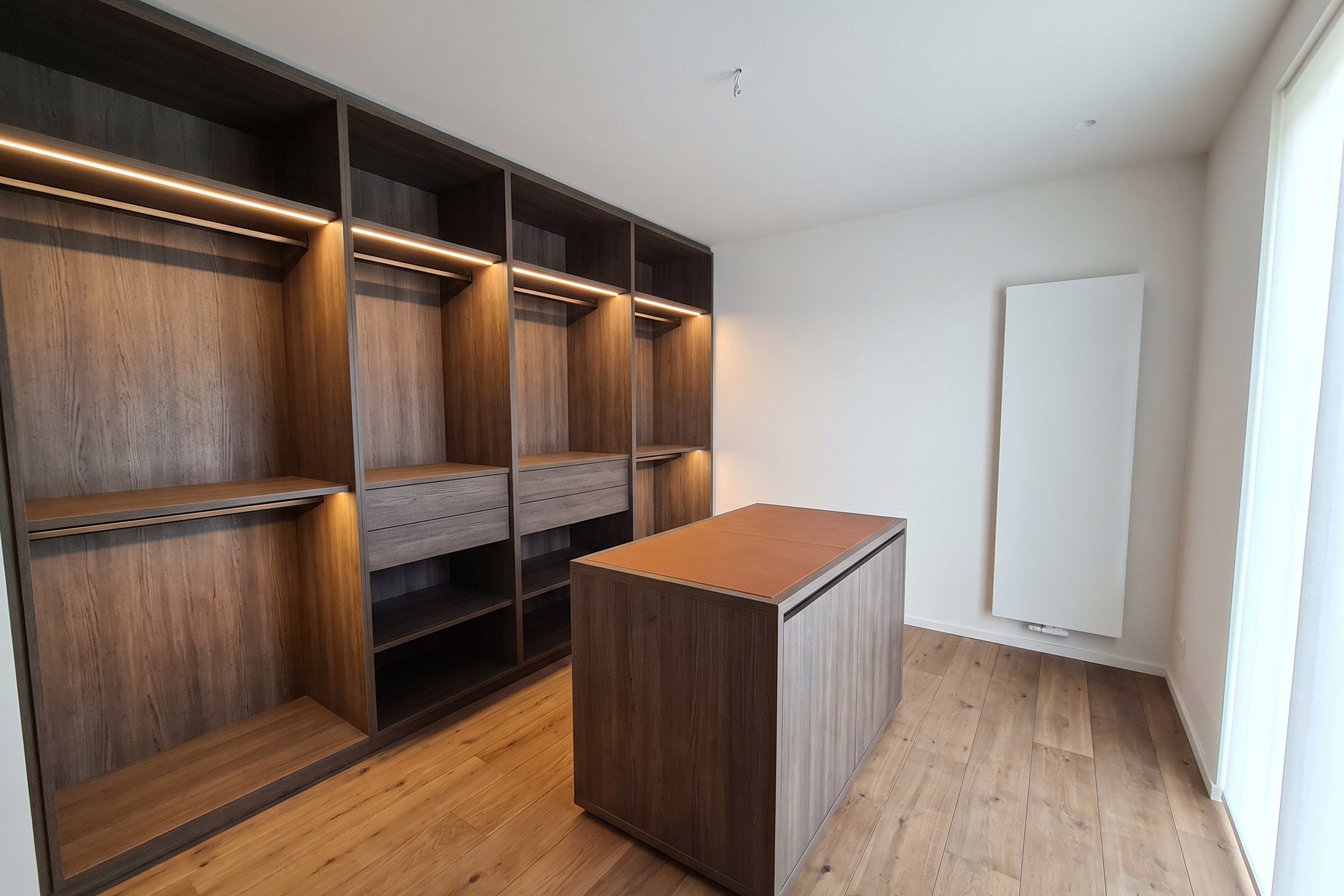 Conception d'un dressing en chêne fumé avec tablettes éclairées, tiroirs et un îlot avec plateau en cuir