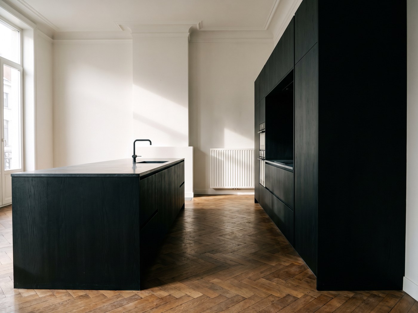 Ontwerp van een keuken in zwart eiken met een eiland en een hoge kastenwand, in een herenhuis met visgraatparket